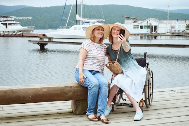 Woman in a wheelchair taking a selfie with her friend against the background of the pier with yachts, catamarans