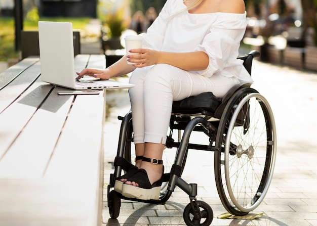 Woman in wheelchair using laptop outdoors while having a drink