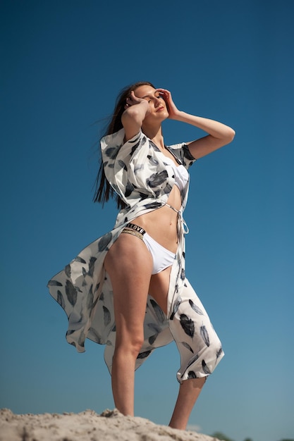 A woman in a white and black swimsuit stands on a beach with her hands on her head.