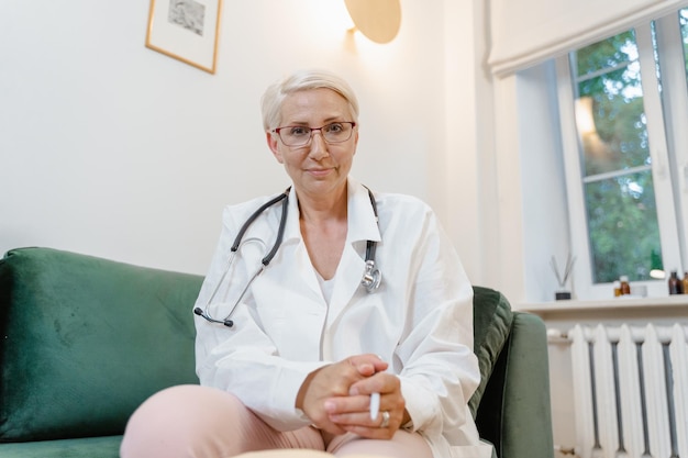 A woman in a white coat sits on a green couch with a stethoscope around her neck.