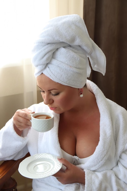 Woman in a white coat and a towel on her head drinks coffee