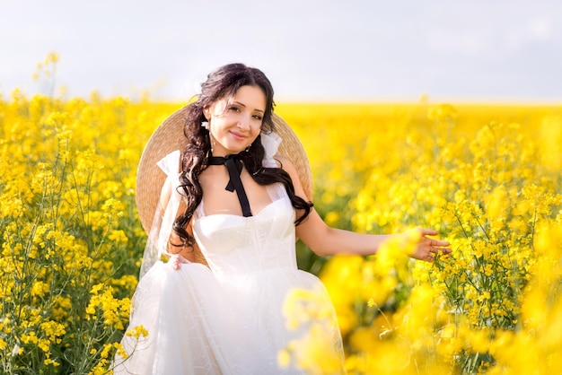 Woman in a white dress and a big hat runs through a rapeseed field Portrait of a girl in yellow flowers on a blue sky background