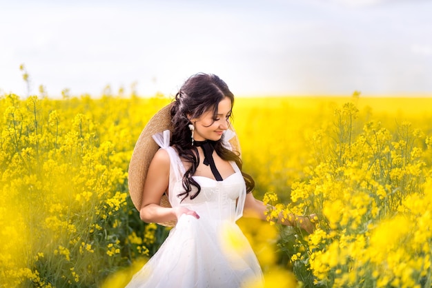 Woman in a white dress and a big hat walks through a rapeseed field Portrait of a girl picking yellow flowers