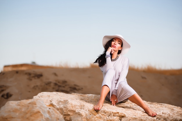 Woman in white dress sitting on a rock at the beach and holding white hat.