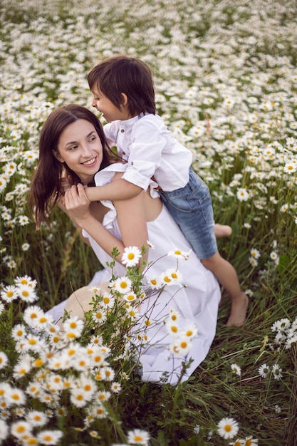 Woman in a white dress with her son a boy of 5 years old lies in a chamomile field at sunset in summer