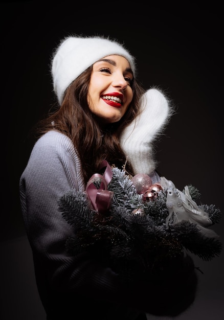 Photo woman in white hat holding christmas decorations a woman wearing a white hat and a purple sweater smiles as she holds a bouquet of fir branches with christmas ornaments