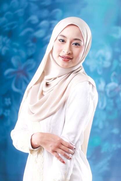 A woman in a white hijab stands in front of a blue background.