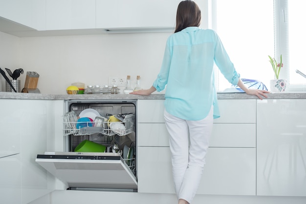 woman in white jeans and shirt stands with her back next to an open dishwasher in kitchen interior