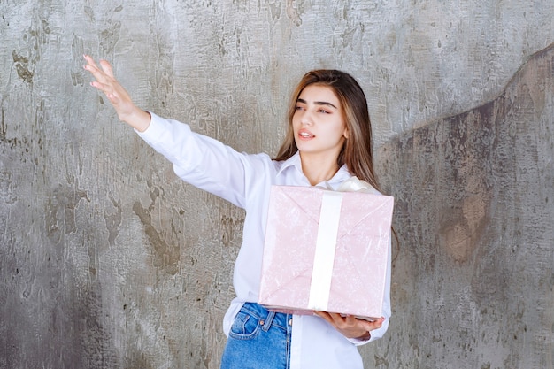 woman in white shirt holding a pink gift box wrapped with white ribbon, noticing her partner and asking him to come and receive it.