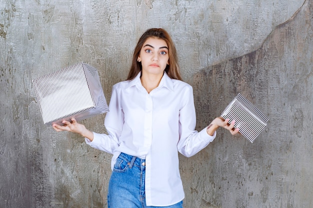 woman in white shirt holding silver gift boxes and looks confused.