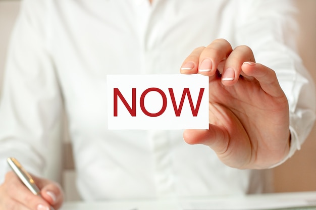 A woman in a white shirt holds a piece of paper with the text: NOW.