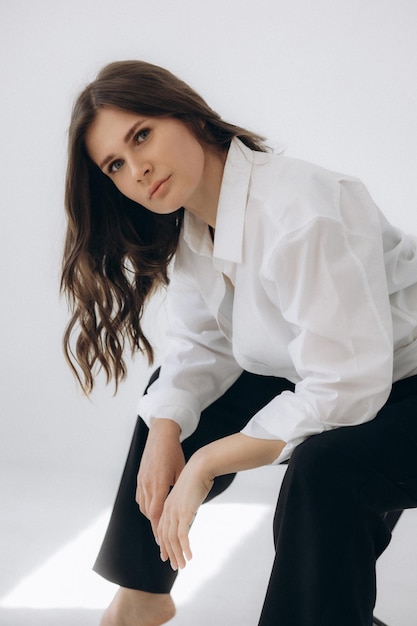 A woman in a white shirt sits on a white surface, wearing black pants and a white shirt.