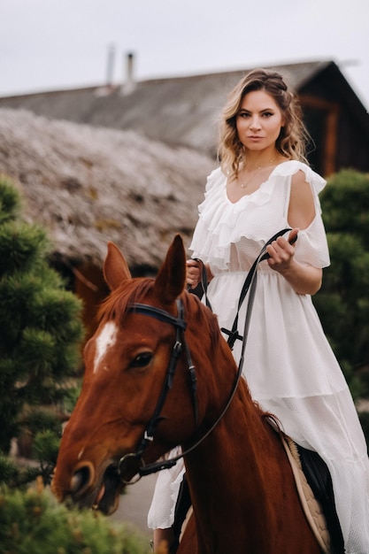 A woman in a white sundress riding a horse near a farm