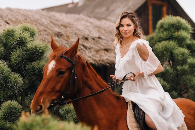 A woman in a white sundress riding a horse near a farm