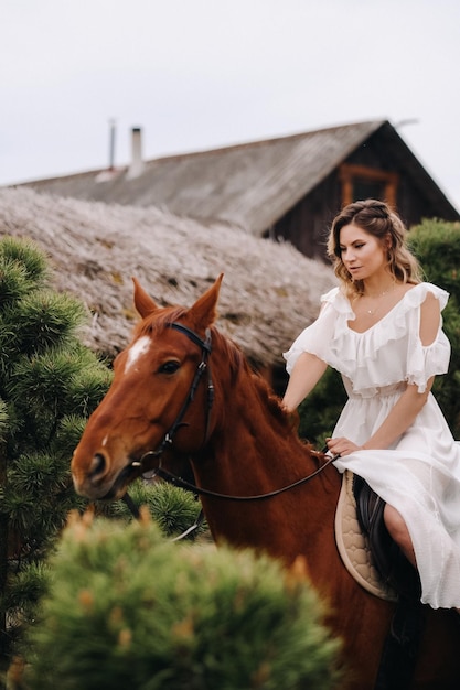 A woman in a white sundress riding a horse near a farm