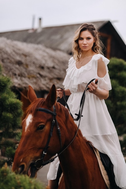 A woman in a white sundress riding a horse near a farm