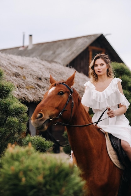 A woman in a white sundress riding a horse near a farm