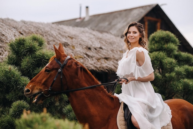 A woman in a white sundress riding a horse near a farm