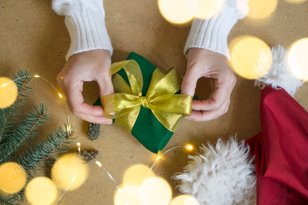 A woman in a white sweater holds a Christmas present on a wooden table background. Atmospheric photo with bokeh lights. Selective focus. Concept of gifts for Christmas and New Year.