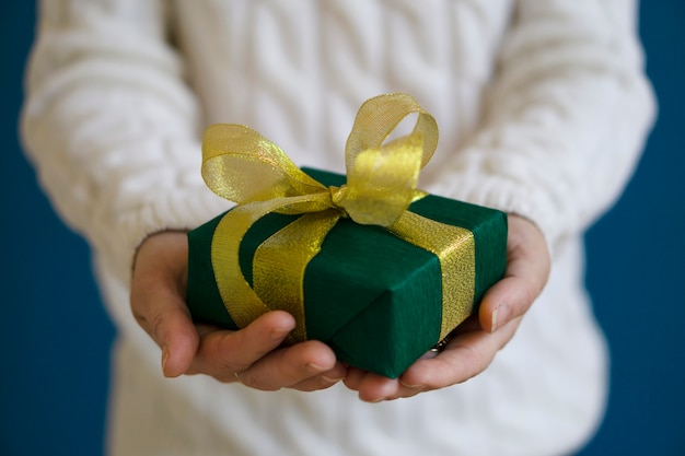 A woman in a white sweater is holding a green Christmas present on a blue background. Gift concept for Christmas and New Year.