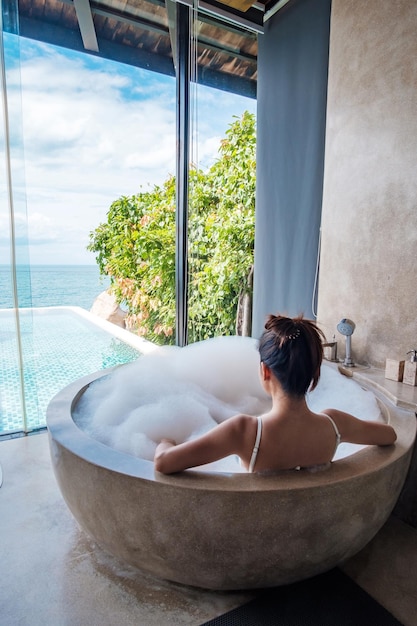 Woman in white swimsuit relaxing in the bathtub with bubble foam against ocean background Summer relaxing and vacation concepts