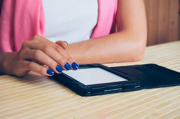 Woman in white T-shirt with a pink scarf and blue manicure reading an e-book at a wooden table