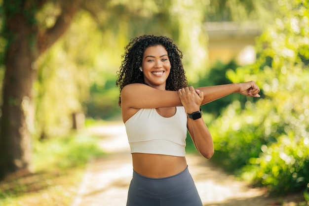 A woman in a white tank top and grey leggings stretches her arm while smiling at the camera in a