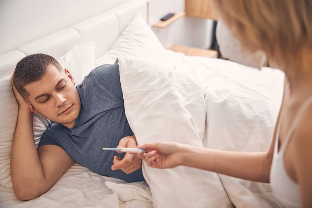 Woman in white top giving thermometer to brunette male while he lying in bed under white blanket