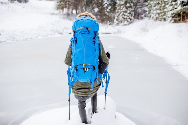 Woman in winter clothes with backpack and tracking sticks standing near the frozen lake during the winter hiking in the mountains