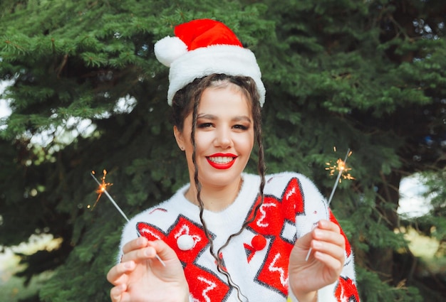 A woman in a winter sweater with a Santa Claus hat and red lips holds a sparkler against the background of Christmas trees new year christmas