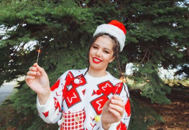 A woman in a winter sweater with a Santa Claus hat and red lips holds a sparkler against the background of Christmas trees new year christmas