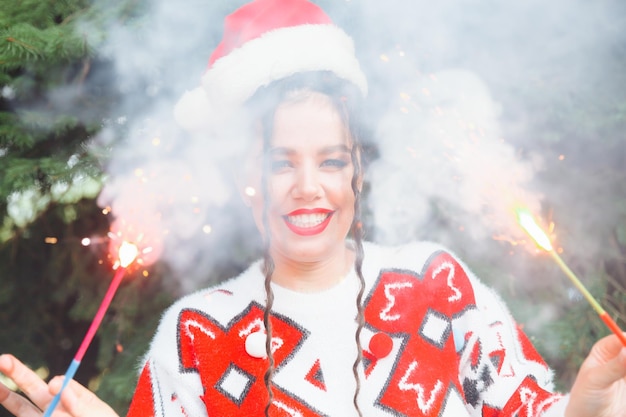 A woman in a winter sweater with a Santa Claus hat and red lips holds a sparkler against the background of Christmas trees new year christmas