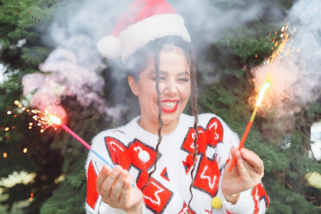 A woman in a winter sweater with a Santa Claus hat and red lips holds a sparkler against the background of Christmas trees new year christmas