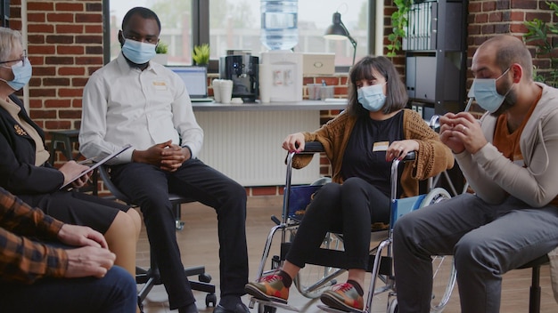 Woman with addiction sitting in wheelchair at aa meeting to do grpup therapy with psychiatrist. Adult with disability wearing face mask and talking about mental health problems.