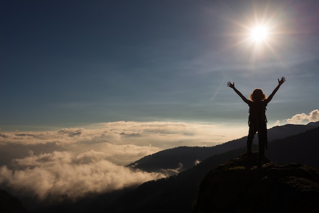 Woman with arms lifted to the sun on mountain top, dramatic landscape clouds over the valley.