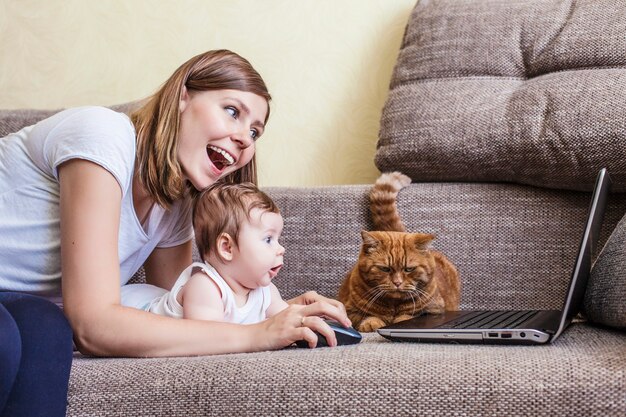 The woman with a baby and a cat at the laptop lying on the couch