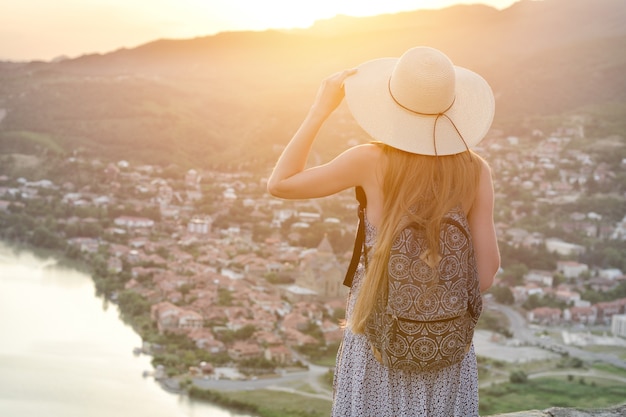 woman with a backpack and in a hat looks at the city and the river below.