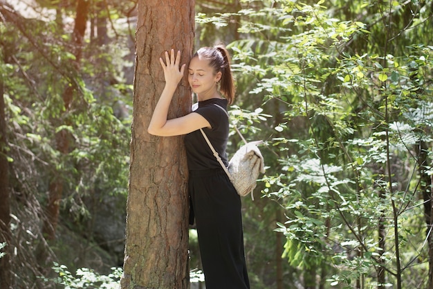 woman with a backpack is hugging a tree with her eyes closed in the forest. Summer sunny day.