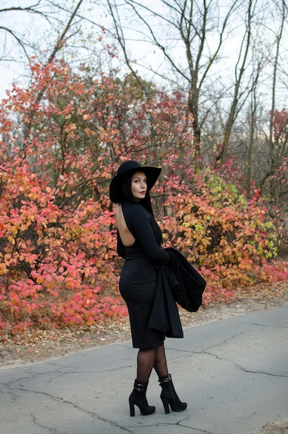 A woman with a beautiful figure in a black hat walks in the autumn park in fulllength