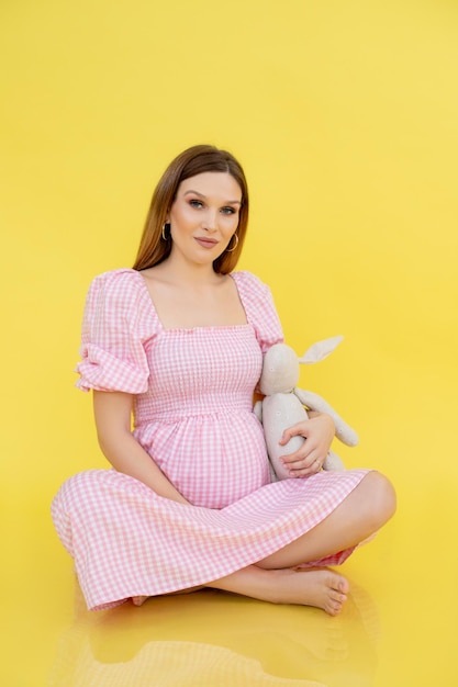 Woman with belly in late pregnancy sit on floor in casual clothes and holding soft toy yellow backgr