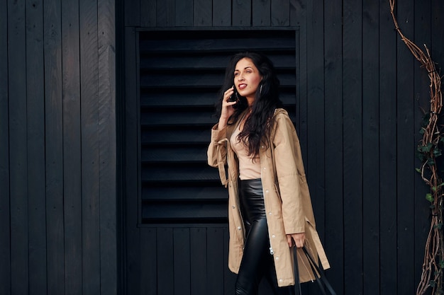 Woman with black curly hair standing against black wooden building exterior and using phone