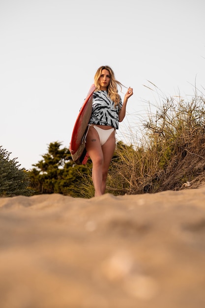 A woman with blonde hair walking on the sand of the beach with a surfboard in summer