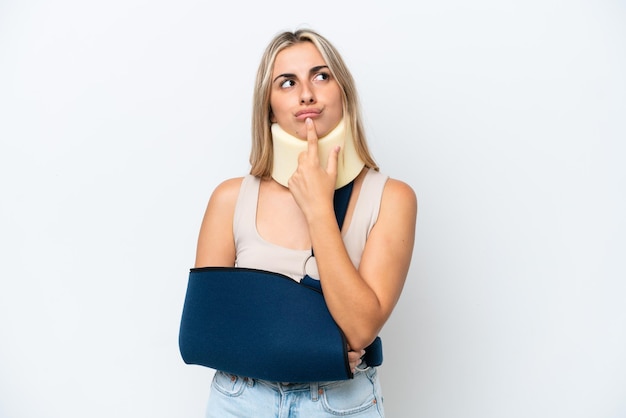 Woman with broken arm and wearing a sling isolated on white background having doubts while looking up