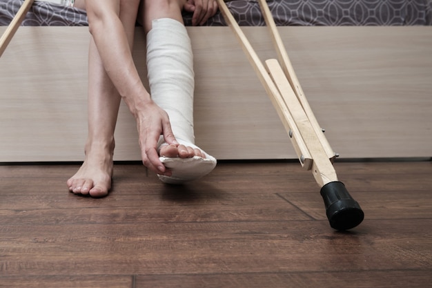 Woman with broken leg sitting on the bed and holding crutches and touch her broken leg