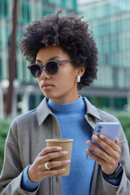 woman with bushy curly hair uses modern digital smartphone for online communication and chatting drinks takeaway coffee wears trendy sunglasses and jacket strolls outdoors in city alone