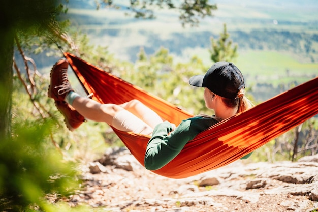 Photo woman with cap resting in comfortable hammock in mountains relaxing on orange hammock between two trees pine enjoying the nature view