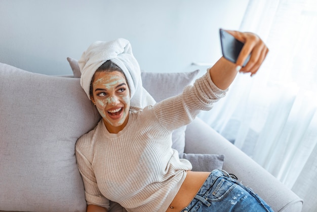 Woman with clay mask taking selfie with mobile phone at home enjoying relaxation