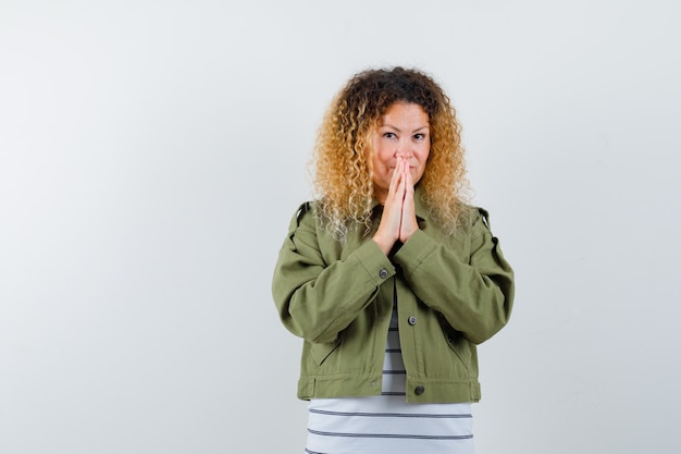 Woman with curly blonde hair in green jacket keeping hands in praying gesture and looking hopeful , front view.