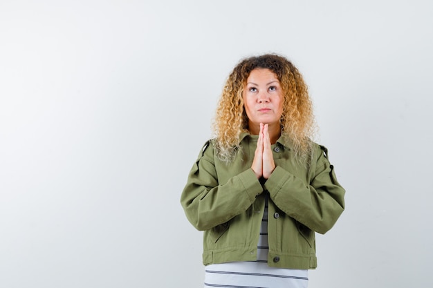 Woman with curly blonde hair in green jacket keeping hands together while praying and looking hopeful , front view.