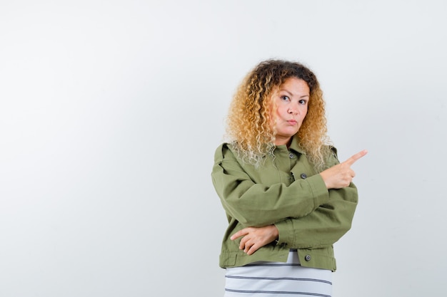 Woman with curly blonde hair pointing at upper right corner in green jacket and looking hesitative. front view.